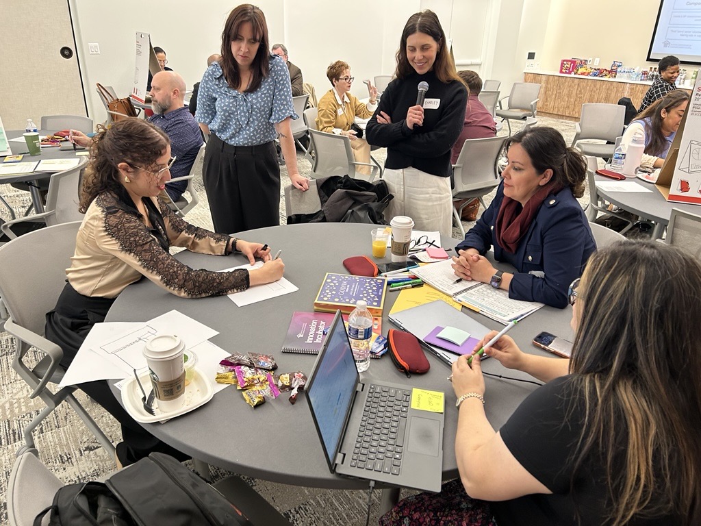 A group of people collaborate around a round table in a bright meeting room. One person sits and sketches on paper while others stand or sit nearby, watching and discussing. The table is covered with notebooks, pens, sticky notes, a laptop, coffee cups, snacks, and a water bottle. In the background, other small groups are also engaged in similar activities, suggesting a workshop or brainstorming session.